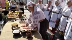 A woman rolling dough balls on a wooden board while other women stand behind and sing