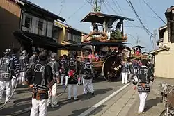 A two-level cart with large wheels and people in festival dress.