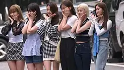 Six women dressed in casual outfits standing in a row outdoors, each striking a pose with their hands under their chins, resembling a flower.