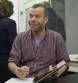Wolfgang Tillmans, a man wearing a checkered shirt. He is looking up, and is holding a pen over a book as if he was going to sign it.