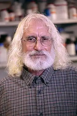 Philip Greenberg wearing a checkered shirt, looking directly at camera, with a blurry background of medications on a shelf