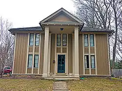 A two-story peaked-roofed wooden house with a projecting front full height porch topped by a pointed roof. It is sided with alternately thick and thin strips of wood running vertically.