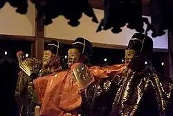 Three masked men in traditional Japanese clothes, one in orange the others in green gold.