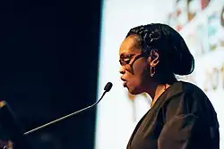 Side profile colour photograph of Nimco Ali speaking from a podium in April 2019.