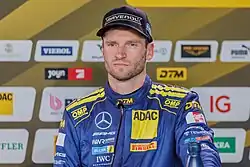 Maro Engel wearing a black baseball cap and blue racing overalls with the logos of sponsors adorning it