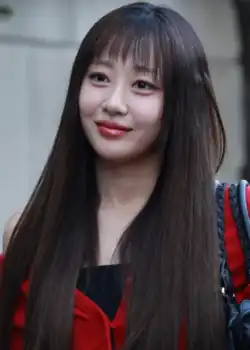 A young Asian woman with long brown hair and a fringe wearing a red off the shoulder cardigan and black sleeveless top carrying a small black leather handbag on her shoulder.