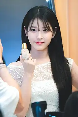 Woman with long, straight black hair wearing a white, off-shoulder dress holding a small bottle with a gold cap in one hand, presenting it to the camera.