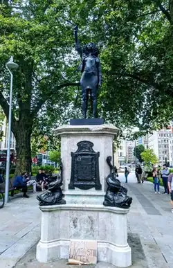 A black life-size statue of a black woman in contemporary clothes, standing with her right fist raised above her head. Statue is on a plinth with a placard placed at the base that reads "BLACK LIVES STILL MATTER". There is a background of an urban space and a tree.