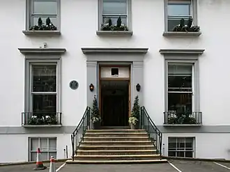 A flight of stone steps leads from an asphalt car park up to the main entrance of a white two-story building. The ground floor has two sash windows, the first floor has three shorter sash windows. Two more windows are visible at basement level. The decorative stonework around the doors and windows is painted grey.