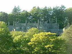 Peeping over the tops of trees in the foreground is a stone house with five gables of different sizes. More trees are in the background.
