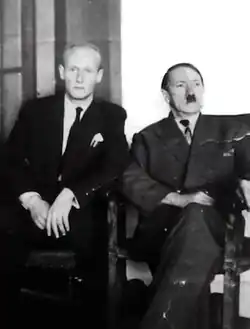 Achromatic, slightly torn photo print of two suited men indoors: to the left sits a balding blond eying the camera and to the right, a sunken-jowled Hitler.