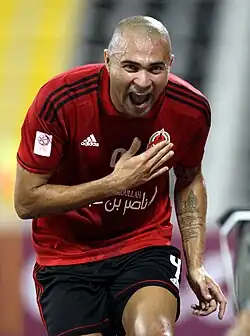 A photograph of a bald Hispanic man in the process of a celebrating a goal, the man is wearing a dark red football shirt and black shorts, he is seen holding his right hand against his chest and with his mouth wide open in the process of cheering.