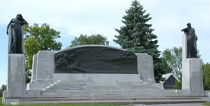 A majestic, broad monument with figures mounted on pedestals to its left and right sides. Along the main portion of the monument are five figures mounted on a broad casting, including a man reclining, plus four floating classical female figures representing Inspiration, Knowledge, Joy, and Sorrow.
