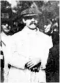 A black and white photo of a man with a handlebar moustache wearing a white jacket and a hat similar to a newsboy cap. He has his hands crossed in front.