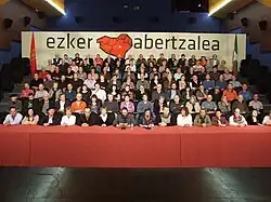 A table with seven steps of people with a banner ezker abertzalea in the background and the flags of Navarre on the left and the Basque Country on the right.