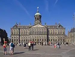 Photo of a symmetrical stone building with a bell tower and triangular pediment with a sculpted relief, with in the foreground a paved square