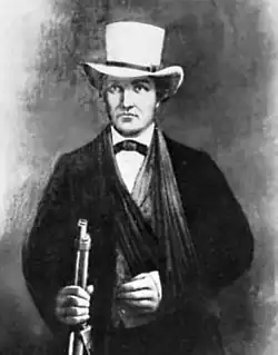 A man in a dark suit with a white hat. His left arm seems to be in a sling. In his right hand he grasps the barrel of a rifle.