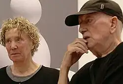 An elderly man and woman dressed in black, with art gallery background. She has dyed, blonde curly hair; he has a baseball cap.