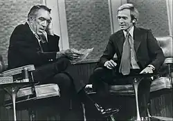 Anthony Quinn and Dick Cavett seated across from one another on a 1971 episode of The Dick Cavett Show. They are both looking at a piece of paper that Quinn is holding; Cavett is smiling.