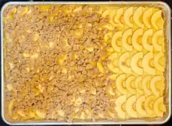 Rectangular baking sheet, with rows of raw apple slices on top of cake batter. Two-thirds of the apples are covered by cinnamon-scented streusel