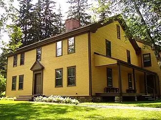 A two-story colonial-style house, painted yellow with brown trim. A porch with benches on it is off the right side of the house.