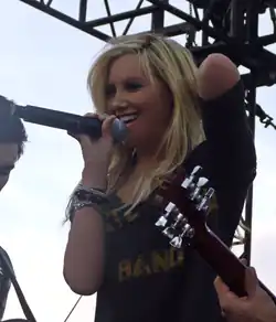 Tisdale, on a stage, sings into a microphone, holding it with her right hand. She is wearing a black blouse with the words "Your" and "Band" printed on it in yellow and few bracelets on her right arm.