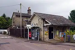 A white-brick station building taken from across the road with a telephone box in front. On the right side of the building there is a door and steps leading down.