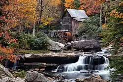 A wooden grist mill standing alongside a rushing stream with a footbridge in the background.