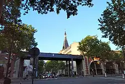 The pedestrian bridge of the Promenade plantée (Coulée verte) traverses the avenue at the intersection of the Avenue Daumesnil. The steeple of St. Antoine Church is also visible.