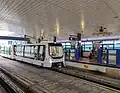 A photograph of Bukit Panjang station's LRT platform, which are two separate platforms facing each other. A train is entering the station, with people at the platform where the train is arriving. The roof is arched and has a series of stripes. There are fans attached to the roof. The platform also feature fixed barriers