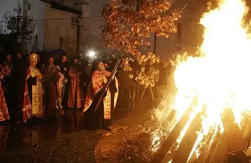 Open-air fire, built with conically arranged long pieces of wood, blazes in the night. Orthodox priest places a long oak sapling with brown leaves on the fire. The priest and the fire are surrounded by a ring of people watching them. In the background, walls of a great church are visible.