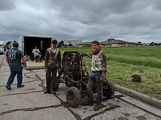 Baja SAE vehicle and two drivers, all completely covered in mud. Ominous rainclouds loom in the background, as well as vehicle trailers and a field of waving grass.