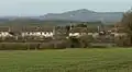 White painted houses with red roofs. In the background is a hill with a tower on it and in the foreground grass and hedgerows