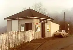 A sepia photo of a very small concrete building viewed from down the road. It has a fence on one side and wall on the other, with a car and a bus stop inside. A sign above the door reads "British Railways / BAYFORD"