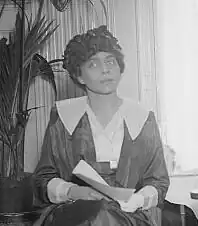 Woman in a dark hat holding a book sits in front of a fronded plant. She is in a white collared shirt, and her head is slightly cocked