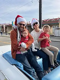 Ben Allen and his wife and two children, sitting in a white convertible car, smiling, wearing Santa hats; outdoors, on a sunny day