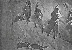 A black-and-white image showing a group of people climbing a fake mountain pass; a body lies on the ground.