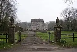fenced and gated driveway leading through grassy area to large two and four storey building in the distance