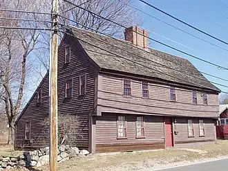 A brown garrison-style saltbox house with a wooden roof and a red door. A rough stone wall separates the street from the yard.