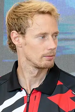 Colour photograph of a man looking to camera-right. He has blond hair, a beard, and is wearing the black, white, and red coloured team jacket of Toyota Gazoo Racing.