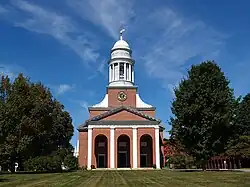 The front of a brick church with white trim. There are three arched openings, lined on each side with white pillar-like facings. The tower has a pillared section in which the church bell is visible, and it is topped by a cupola and a weathervane.