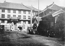 A black and white image of several buildings with Chinese-style wide-eaved roofs making a corner around a parking area where a 1920s-style automobile is parked