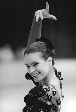 An image of Katarina Witt smiling and throwing up her right hand with the index finger pointed upwards and the other fingers parallel to the ground.