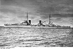 A large gray warship with four large gun turrets and two tall funnels sits idly in harbor.