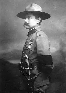 Photo of Burnham taken in 1901 in London after his investiture with the cross of the Distinguished Service Order by King Edward VII. He is dressed in British Army uniform and standing at attention, facing right. On his left arm is a black armband worn in mourning for the recent death of Queen Victoria. He is wearing his stetson hat and a sword on his left side. He sports a large handlebar mustache.
