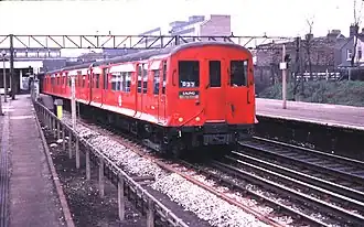 A three-quarter photograph of a red train O stock with sliding doors and flared sides, leaving Barking station bound for Ealing in 1980