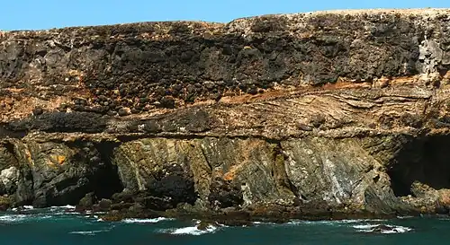 Cliff at Caleta Negra, Ajuy on Fuerteventura. The lower third of the cliff consists of steeply-tilted pre-Miocene oceanic sedimentary rocks. These rocks are unconformably overlain by Pliocene light-coloured cross-bedded beach sedimentary rocks, black pillow lava, and a black lava flow.[301][302] (Scale: cliff height is approximately 40 m (130 ft)[303])
