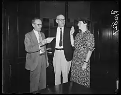 Two white men and a white woman, all standing; the woman has her hand raised in a position of swearing an oath; the woman is wearing a print dress, and the men are wearing suit jackets and ties