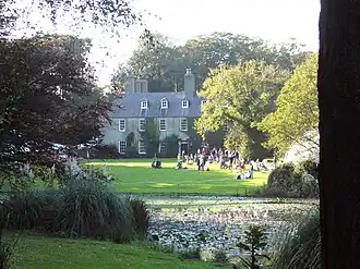 a house with trees to each side and a lawn in front of it, there is a lake in the foreground