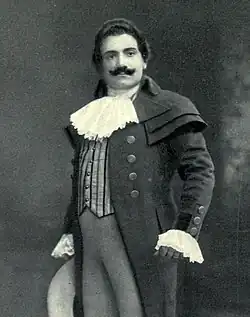 Man, with dark hair and a curling moustache, standing in a posed position. He is wearing a long coat, with lace at the throat and cuffs.